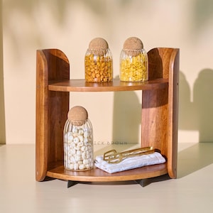 Puede incluir: Una estantería de madera de dos niveles con tres tarros de cristal llenos de aperitivos y tapados con tapas de corcho. Un utensilio de servir de color dorado descansa sobre un paño blanco y beige doblado en el estante inferior. La estantería es de madera de tonos cálidos.