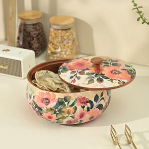 May include: A floral-patterned wooden bowl with a matching lid, partially open to reveal tortillas. The lid and bowl feature a cream background with pink and orange flowers and green leaves. Two glass jars with wooden lids are in the background.