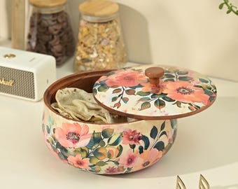 Puede incluir: Un recipiente redondo de madera con un estampado floral de flores rosas y naranjas y hojas verdes. El recipiente tiene una tapa a juego con un pequeño pomo y está lleno de panes planos o galletas saladas finas y claras.
