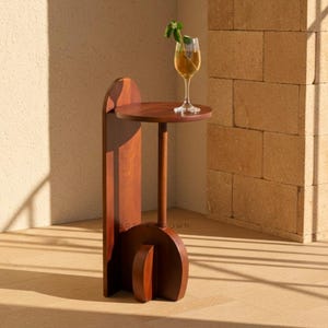 May include: A wooden side table with a round top and a tall, slender stem. The table is made of dark wood and has a unique, geometric base. A glass of liquid with a sprig of greenery sits on top.