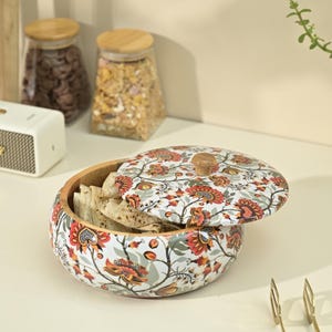 May include: A round wooden bowl with a floral design lid, filled with flatbread. The bowl's exterior features a white background with red, orange, and green floral patterns. The lid has a wooden knob. The bowl is approximately 8 inches in diameter.