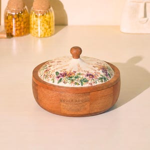 May include: A round wooden bowl with a floral patterned lid. The lid features a cream background with purple, pink, and green flowers. The bowl is made of light brown wood and has a small wooden knob on top.