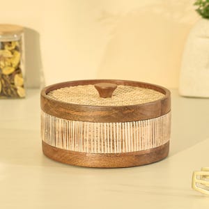 May include: A round wooden storage box with a woven rattan lid and a band of vertical rattan strips around the middle. The box has a small, rounded wooden knob on top. The wood is a warm brown color.
