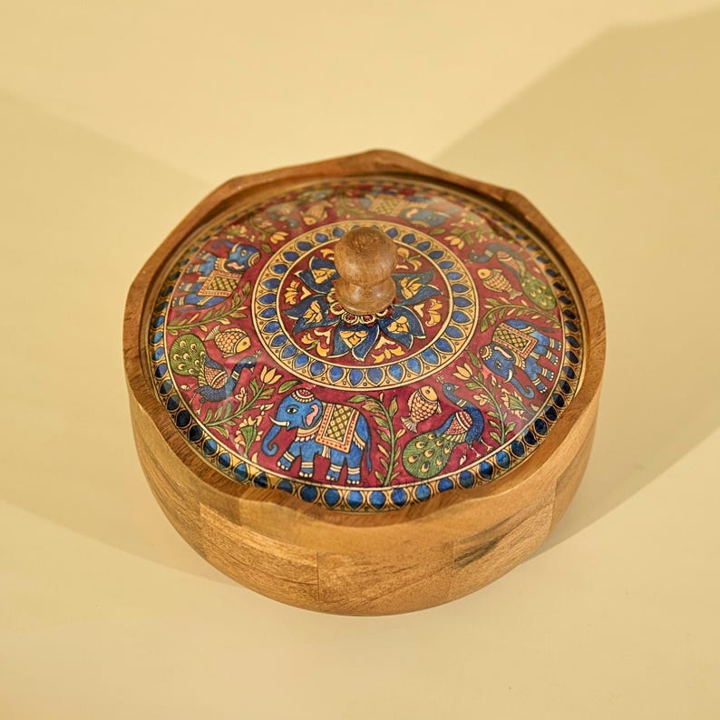 May include: A wooden, hexagonal-shaped box with a lid. The lid is adorned with a detailed, colorful design featuring elephants, peacocks, and floral patterns on a maroon background. The box has a natural wood finish.