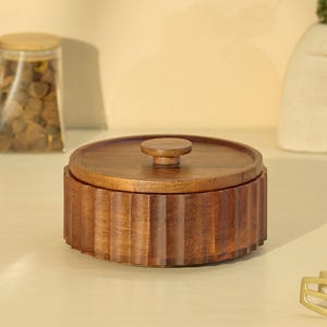 May include: A round, wooden container with a lid and a small knob. The container has a ribbed design around the sides and is made of a warm, brown wood. The lid fits snugly on top. The container is likely used for storage.