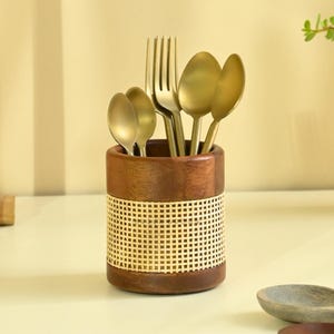 Könnte beinhalten: Ein Holz-Utensilienhalter mit einem gewebten Rattanband, der goldfarbene Löffel und eine Gabel enthält. Der Halter ist zylindrisch und steht auf einer hellen Oberfläche. Die Löffel und die Gabel sind dekorativ angeordnet.