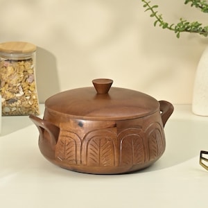 May include: A wooden cooking pot with a lid and two handles. The pot is brown and has carved leaf designs around the base. The lid has a small, rounded knob. A glass jar with a wooden lid is in the background.