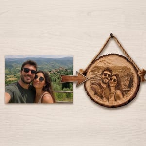 Puede incluir: Una fotografía rectangular en color de una pareja con gafas de sol al aire libre con un paisaje montañoso de fondo, conectada por una flecha de madera a una placa circular de madera con una versión grabada a láser de la misma imagen.