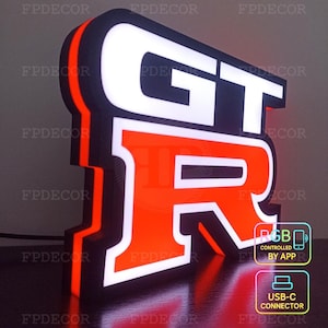 Op de afbeelding: Verlicht bord met de letters "GT-R" in wit en oranje, met een zwarte rand. Het bord is verlicht met RGB-lampen en wordt bestuurd via een app. Het bevat een USB-C-connector.