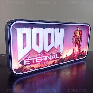 May include: Rectangular LED light box featuring the "DOOM Eternal" logo and a character illustration. The design has a black frame with a red and purple background. The text "DOOM" is in large white letters, with "ETERNAL" below.