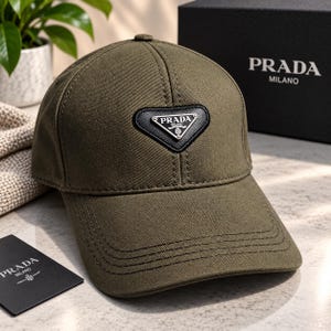 Pode incluir: Um boné de beisebol verde-oliva com um emblema triangular de couro preto na frente com o logotipo Prada Milano. O boné possui uma coroa estruturada, aba curva com costuras tonais e é acompanhado por uma caixa preta da marca Prada Milano.