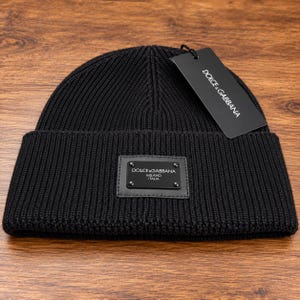 Puede incluir: Un gorro de punto acanalado negro con una placa metálica rectangular negra en la parte frontal con el texto DOLCE&GABBANA MILANO ITALIA. Una etiqueta de papel rectangular negra está sujeta en la parte superior, mostrando DOLCE&GABBANA.