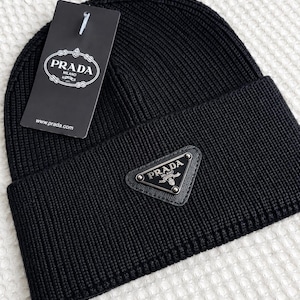 Op de afbeelding: Een zwarte, geribbelde gebreide muts met een driehoekig metalen Prada Milano-logoplaatje op de voorste omslag. Een zwart rechthoekig Prada Milano-merklabel is aan de bovenkant van de muts bevestigd met een witte plastic sluiting.