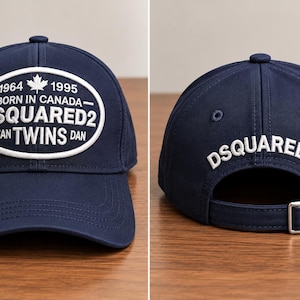 May include: A navy blue baseball cap featuring a white oval patch on the front with the text 1964 1995 BORN IN CANADA DSQUARED2 DEAN TWINS DAN and a maple leaf. The back displays the white embroidered text DSQUARED2 and an adjustable metal buckle.