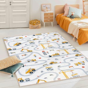 May include: A rectangular children's rug with a construction theme. The rug features a white background with a road design and illustrations of yellow construction vehicles, buildings, and cranes. The rug is bordered with a light beige trim.
