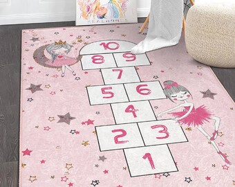 May include: Pink play rug with a hopscotch game and two ballerina figures. The hopscotch game has numbers 1 through 10. The rug is covered in small pink stars.
