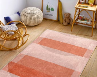 May include: A pink and orange striped area rug with a subtle textured pattern. The rug is laid out on a wooden floor in a room with a rocking chair, a stuffed animal, and a picture frame with the text "Real Homes".