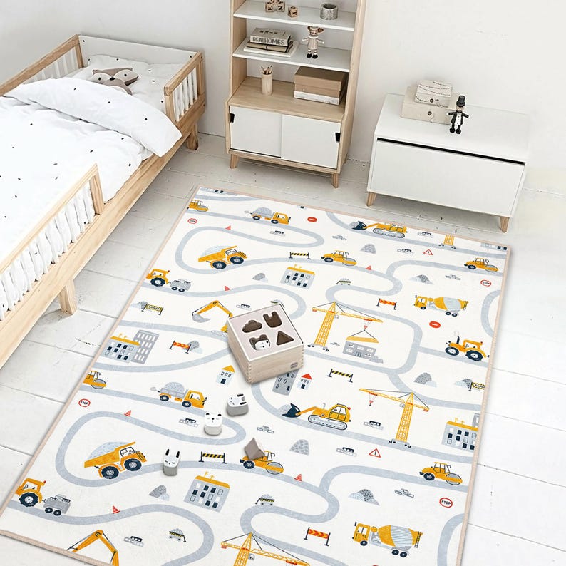 May include: A children's play mat with a road and construction vehicle design. The mat features yellow trucks, cranes, and buildings on a white background with gray roads. A wooden shape sorter and toy blocks are on the mat.