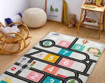 May include: A grey and white play rug with a road and number design. The rug features a black road with a yellow traffic sign, a traffic light, and a stop sign. The numbers 1 through 9 are in colourful squares along the road.