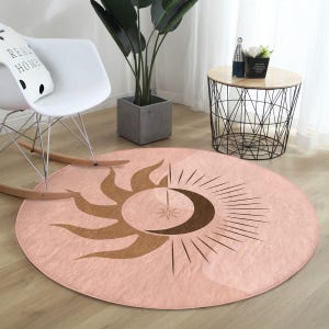 Pode incluir: Um tapete redondo cor de rosa blush com um design celestial. O tapete apresenta um motivo de sol e lua crescente castanho com linhas radiantes e uma estrela central. O tapete está colocado sobre um chão de madeira, com uma cadeira de baloiço branca e uma mesa lateral no fundo.