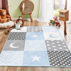 Puede incluir: Una alfombra de juego azul y gris con un diseño de patchwork con estrellas, lunas y lunares. La alfombra es perfecta para una guardería o sala de juegos.