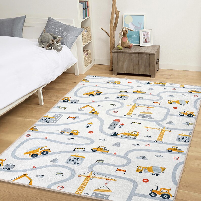 May include: A rectangular children's rug with a construction theme. The rug features a white background with a gray road design and yellow construction vehicles, including trucks, excavators, and cranes. The rug is designed for a child's bedroom or playroom.