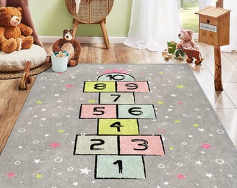 May include: A grey and white play rug with a hopscotch design. The hopscotch squares are numbered 1 through 10 and are coloured pink, yellow, and green. The rug is decorated with white stars and circles.