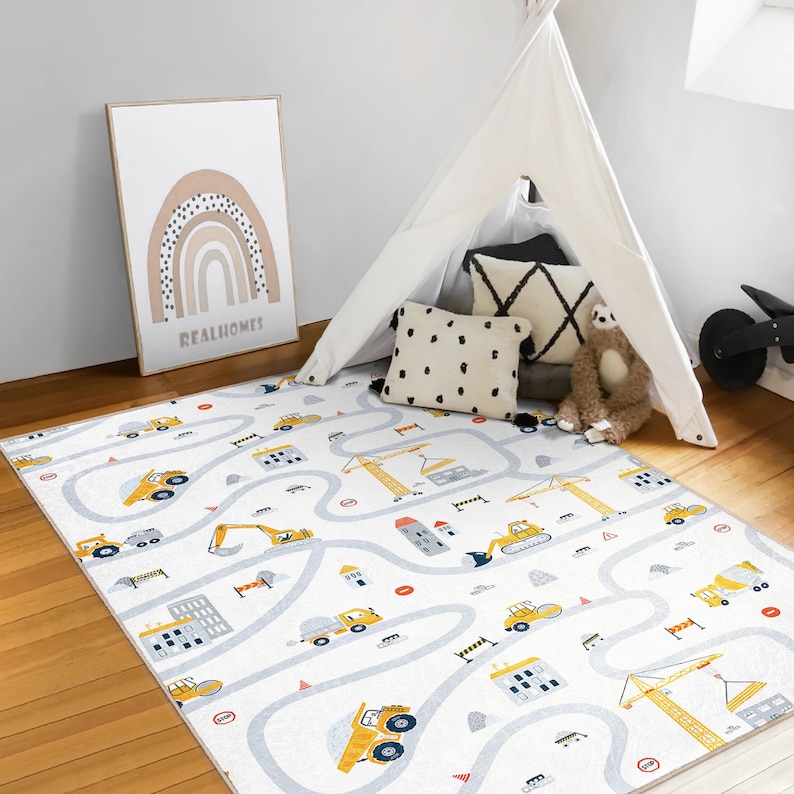 May include: A white play mat with a road and construction vehicle design. The mat features yellow construction vehicles, buildings, and cranes on a winding gray road. A framed rainbow art print and a white teepee are in the background.