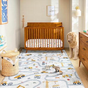 May include: A nursery scene featuring a wooden crib, a road-themed rug with construction vehicles, and a woven basket. The rug has a light gray background with a road design and yellow construction vehicles. A stuffed lion stands near a wooden dresser.