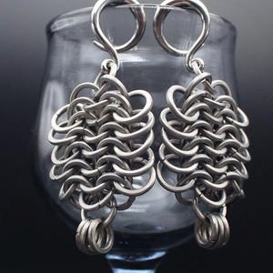 Puede incluir: Un par de pendientes de cota de malla plateados. Cada pendiente presenta una parte superior en bucle para colgar, un diseño de cota de malla tejida y un pequeño grupo de anillos en la parte inferior. El fondo está borroso.