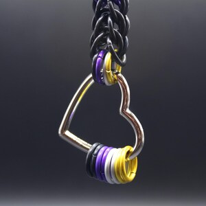 May include: A silver heart-shaped keychain with a chain of black rings. The keychain features rings in purple, yellow, white, and black, representing the non-binary flag. The design is simple and elegant.