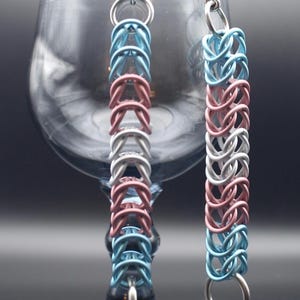 Puede incluir: Un par de pendientes de cota de malla hechos a mano. Los pendientes están hechos de pequeños anillos de metal entrelazados en azul claro, rosa y plateado. Los pendientes cuelgan frente a una copa de vino.