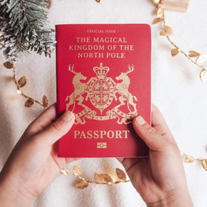 May include: A red passport with gold lettering and a gold emblem is held. The text reads "OFFICIAL ISSUE", "THE MAGICAL KINGDOM OF THE NORTH POLE", and "PASSPORT". A gold garland and a Christmas tree branch are in the background. Festive theme.