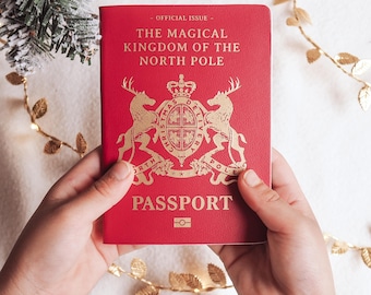 May include: A red passport with gold lettering and a gold emblem is held by two hands. The text on the passport reads "OFFICIAL ISSUE", "THE MAGICAL KINGDOM OF THE NORTH POLE", and "PASSPORT". A gold garland and Christmas tree branch are in the background.