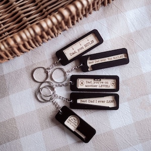 May include: Set of four black wooden keychains with laser-cut designs and sayings. The keychains feature a ruler, hammer, level, and saw. The sayings on the keychains are "Dad, you RULE!", "Best Dad ever. You NAILED it", "Dad, you're on another LEVEL!", and "Best Dad I ever SAWI".