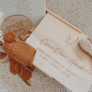 May include: A wooden box with a light brown finish, engraved with the name "Oscar Romio Grech", the date "February 13, Twenty Twenty", the time "11:34PM", the weight "3.02 kg", and the length "49 cm". The box contains a knitted hat, a wooden toy, and a wooden sign that says "earth side".