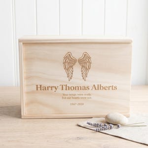 May include: A rectangular light-coloured wooden box featuring an engraved pair of angel wings. The text reads: Harry Thomas Alberts, Your wings were ready, but our hearts were not, 1947-2020. A smooth white stone and a sprig of dried lavender lie nearby.