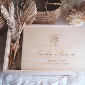 Peut inclure: Un coffret souvenir en bois clair avec le nom "Caeley Rainne" gravé sur le couvercle, ainsi que les détails de naissance. Le couvercle présente un motif floral et lunaire. Le coffret est entouré d'objets décoratifs.
