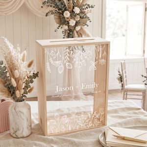 May include: A rectangular wooden card box with a clear acrylic front panel featuring white line art of wildflowers and the text "Jason + Emily" along with "Seventh of November, Two Thousand and Twenty-six" printed in a white serif font.