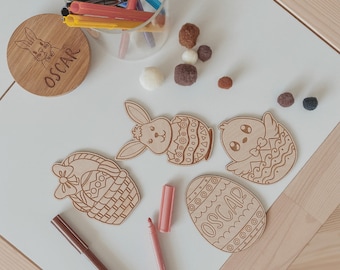 以下が含まれることがあります： 木製のイースターをテーマにした装飾品。名前「OSCAR」が入ったウサギのコースター、ウサギ、ヒヨコ、バスケット、卵のレーザーカットの形。カラーマーカーとフェルトボールも写っています。