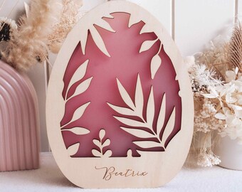 May include: Wooden Easter egg ornament with a cut-out leaf design and a pink background. The word "Beatrix" is written in cursive at the bottom. A white ribbon is attached for hanging. The ornament is surrounded by dried flowers.