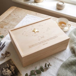 May include: A rectangular light-coloured wooden keepsake box featuring a gold star emblem, engraved constellation patterns, and the text: Dad, The brightest star in the sky shines as a constant beacon of love, a reminder that you are never truly gone, but always watching over us, 1947-2020.