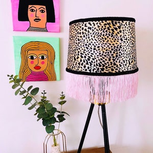 Può includere: Una lampada a treppiede nera con un paralume a stampa leopardata. Il paralume ha un bordo nero e una frangia rosa. La lampada è su un tavolo di legno con un vaso di metallo dorato con una pianta verde.