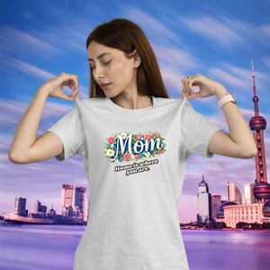 Könnte beinhalten: Weißes T-Shirt mit dem Wort "Mom" in einem floralen Design. Darunter steht der Satz "Home is where you are". Das T-Shirt wird von einer Person präsentiert.