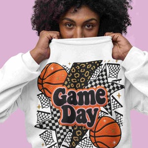 Könnte beinhalten: Weißes Sweatshirt mit Basketball-Motiv. Das Design umfasst orangefarbene Basketbälle, Sterne und den Text "Game Day". Der Satz "Sparkle on the Court" ist ebenfalls sichtbar.