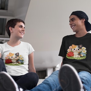 Könnte beinhalten: Zwei T-Shirts, eines weiß und eines schwarz, mit einem farbenfrohen Katzendesign, das von Blumen umgeben ist. Die Katze hat orangefarbenes, weißes und schwarzes Fell und einen Bogen auf dem Kopf. Die Blumen umfassen rote Rosen und blaue und weiße Gänseblümchen.