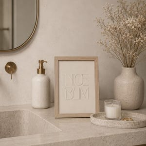 Op de afbeelding: Een rechthoekige lijst van licht hout met een crèmekleurige print met de in reliëf aangebrachte tekst NICE BUM in een strak, schreefloos lettertype. De lijst staat rechtop op een aanrecht en toont een duidelijke en leesbare tekst.