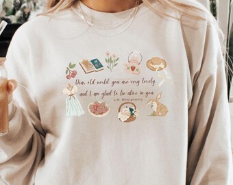 May include: A beige sweatshirt with a quote from L.M. Montgomery printed on the front. The quote reads "Then old world you are very lovely and I am glad to live in you". The quote is surrounded by small illustrations of flowers, a book, a hat, and a rabbit.