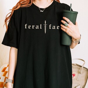 Puede incluir: Camiseta negra con el texto "feral fae" en fuente gótica, con un gráfico de espada. Se sostiene un vaso verde oscuro en la mano. El fondo incluye una mochila y una manta.