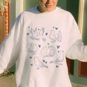 May include: White sweatshirt with a blue graphic of multiple pigeons and hearts. The person wearing the sweatshirt is holding a cup of coffee.